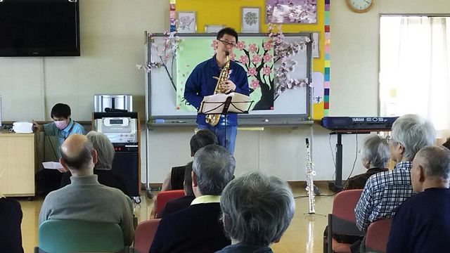 桜の季節の音楽祭「春の宴」での、サックス奏者の演奏と聞く高齢者